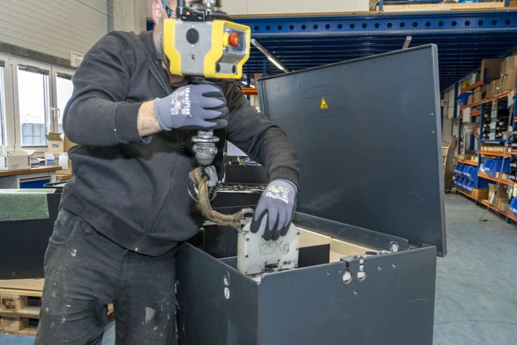 An employee at AKKU SYS uses a crane module to lift a 125 kg battery element into an open black battery housing.