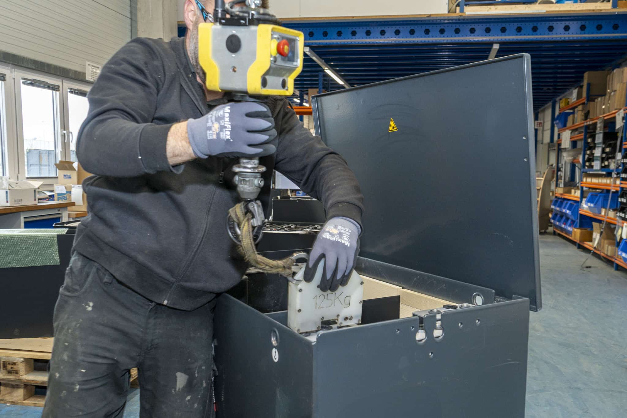 An employee at AKKU SYS uses a crane module to lift a 125 kg battery element into an open black battery housing.