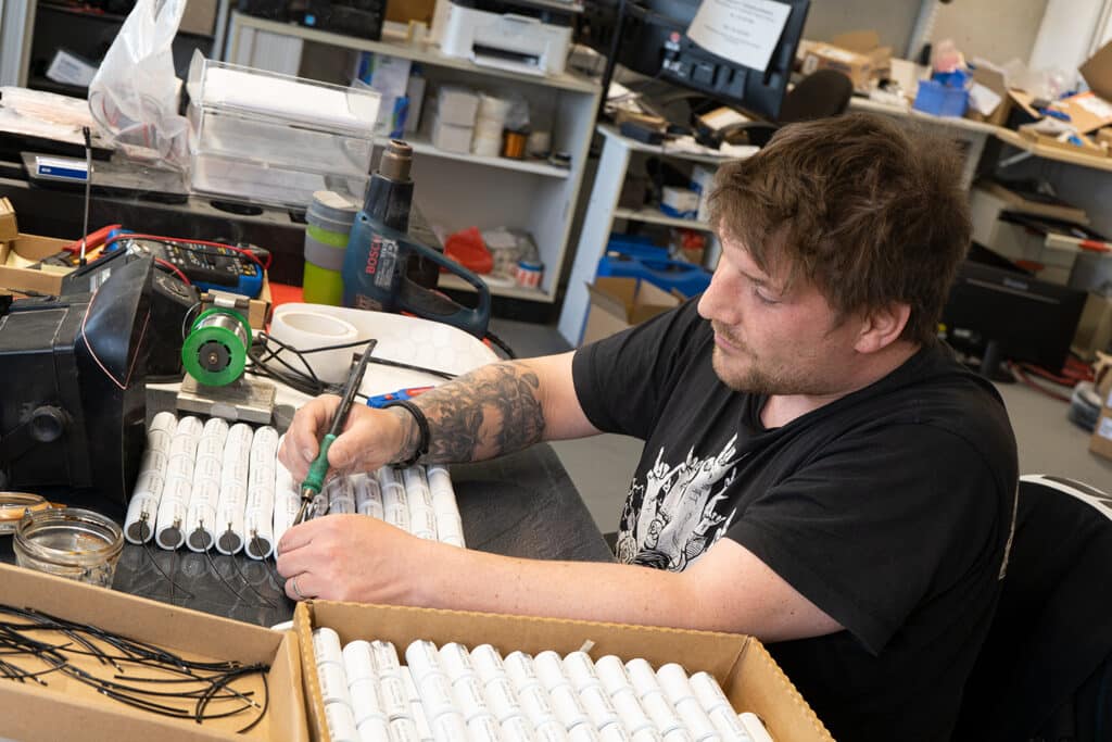 AKKU SYS employee in a workshop soldering wire connections to cylindrical battery cells that lie in several rows in front of him.