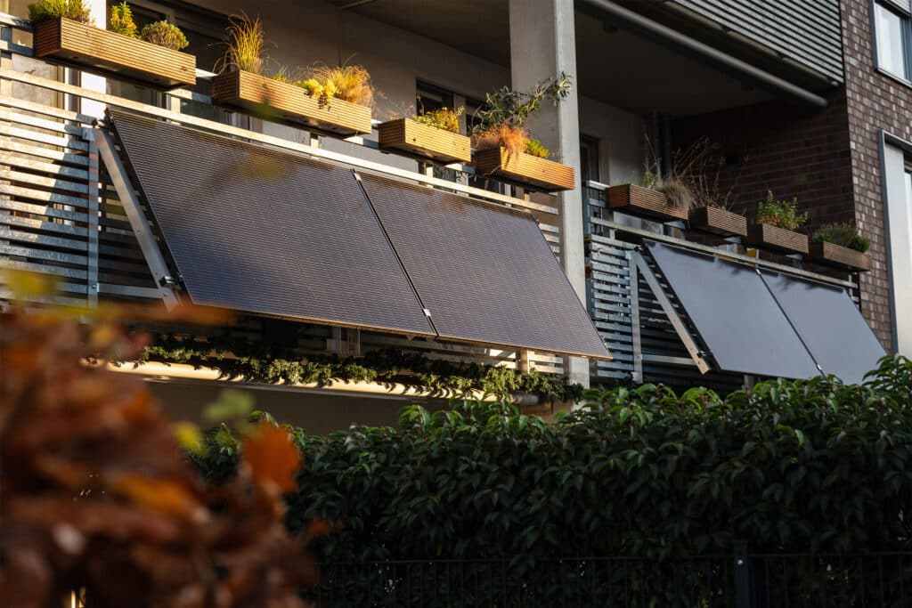 Balcony power station with two solar panels mounted on the parapet of an apartment block balcony. Above the solar panels are planter boxes with grasses and small plants. Other balconies with similar systems and planter boxes can be seen in the background, with a bush in the foreground.