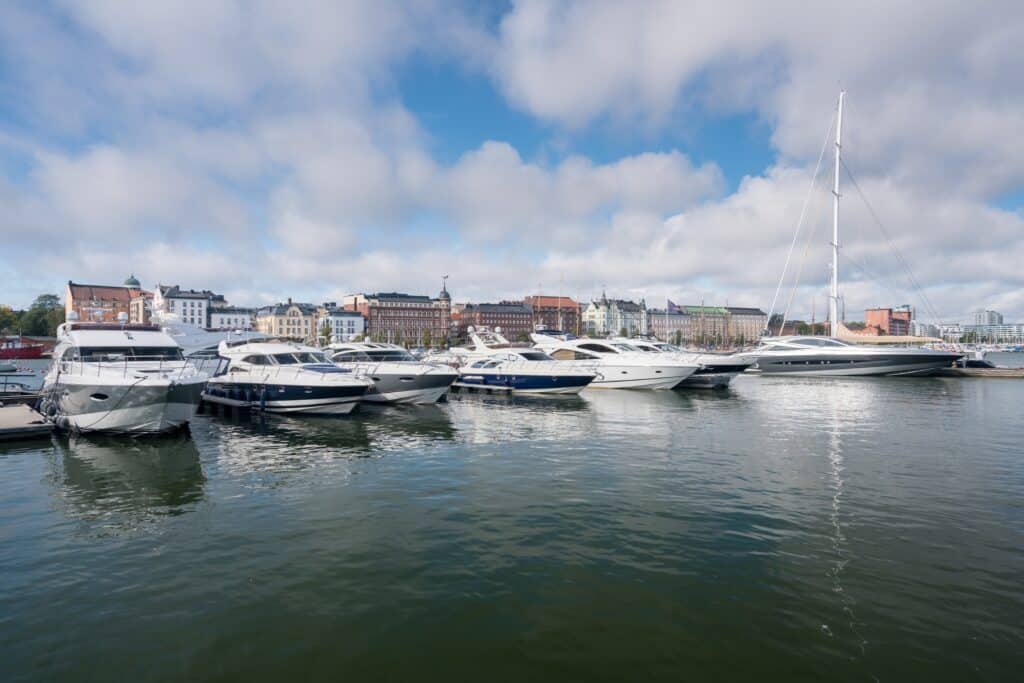 Mehrere Motorboote und eine große Segelyacht liegen in einem Hafen vor einer Stadtkulisse mit historischen Gebäuden unter blauem Himmel mit Wolken.