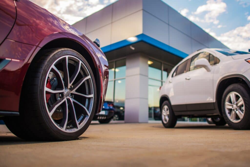Close-up of a red sports car wheel in front of a white SUV in the car park of a car dealership with a modern glass façade and blue canopy.