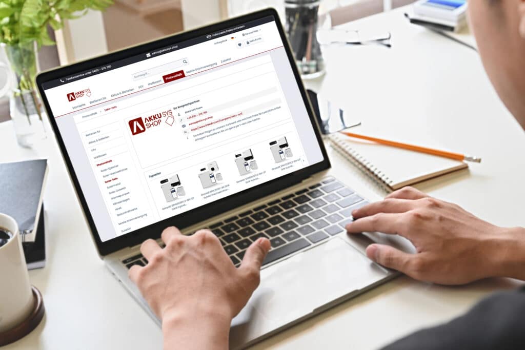 Person uses a laptop and visits the AKKU SYS online shop. The screen shows various products for mobile power supplies and photovoltaic components.