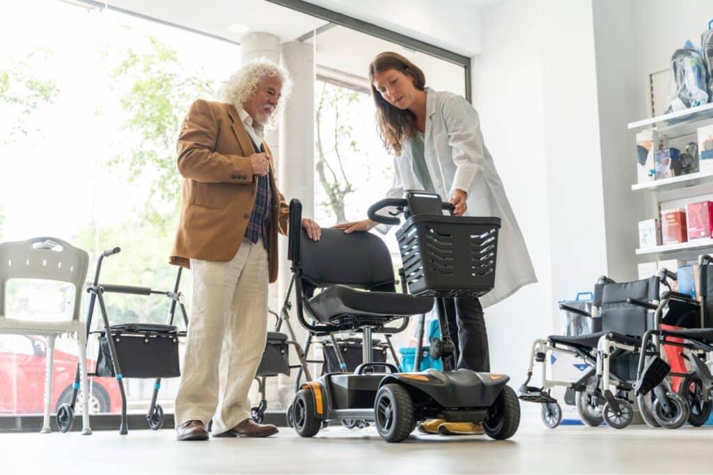 Consultation in a specialist rehabilitation shop, where an employee demonstrates a mobility scooter to an elderly man. Wheelchairs and walking aids can be seen in the background.