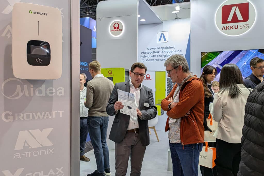 AKKU SYS exhibition stand at a solar technology trade fair. An employee explains technical details to a visitor using a flyer. Other visitors, exhibition stands and brand logos such as Midea, Growatt and a-TroniX can be seen in the background.