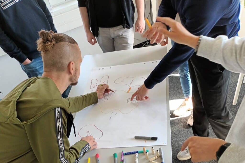 A large sheet of paper and different coloured pens lie on a table. Five people stand around the table and work together on a sketch.