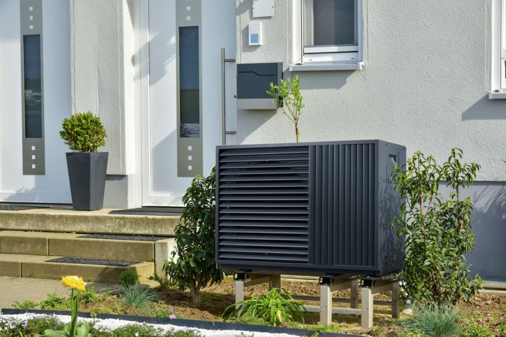 A black air-to-water heat pump, installed in front of the white façade of a private home.