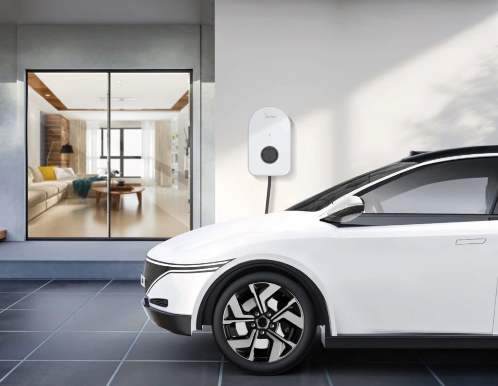White electric car on a Midea wallbox in front of a modern residential building with a view into the living room.