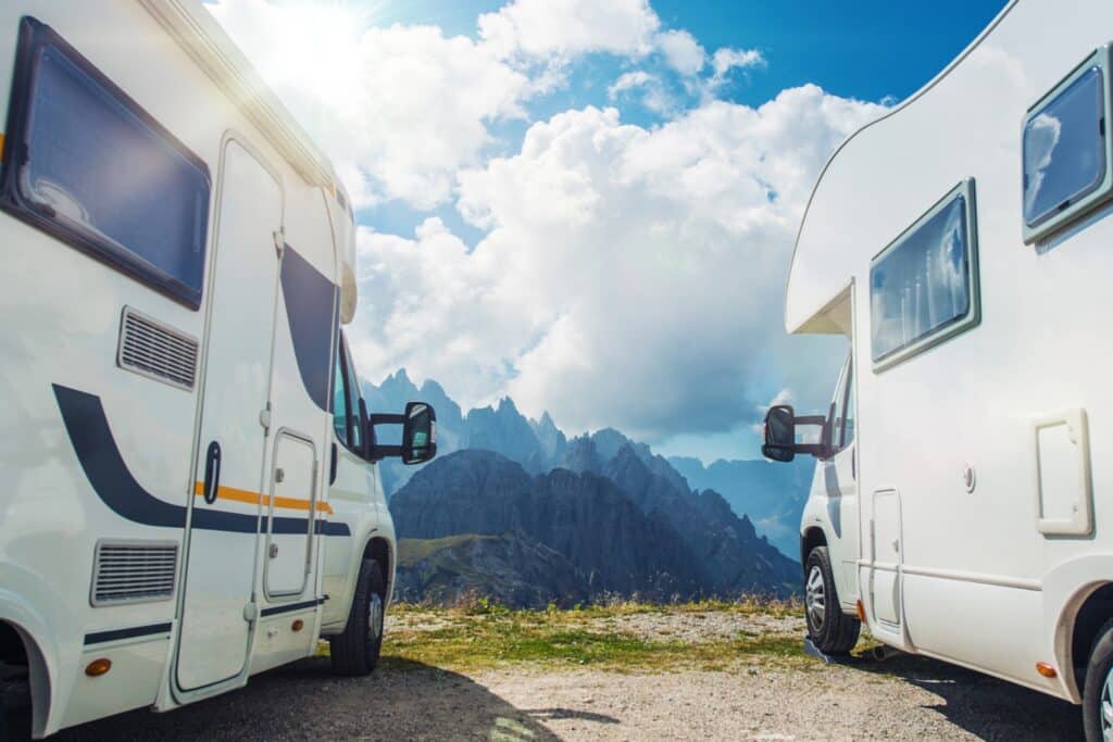 Two motorhomes are parked on a mountain plateau with a view of a rocky mountain landscape under a blue sky with clouds.
